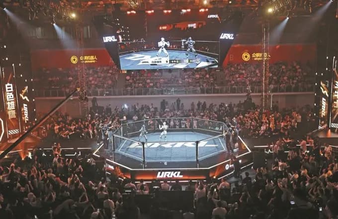 Público alrededor de una jaula octogonal con el rótulo URKL, dos robots humanoides en su interior y una pantalla del combate.